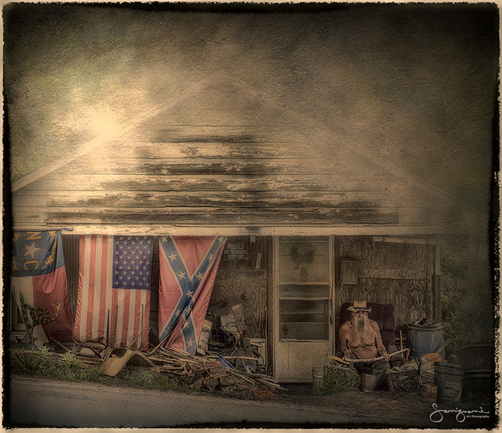 weathered cabin with nc, us, and confederate flags. shirtless man whittling wood sits on porch.